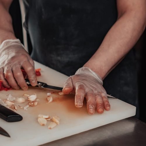 A chef wearing gloves slices garlic and red pepper on a chopping board, showcasing precision and hygiene.